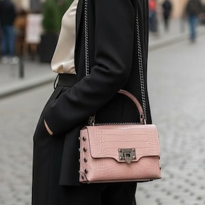 Puede incluir: Una mujer con blazer y pantalón negros está de pie en una calle adoquinada, sosteniendo un bolso rosa claro con una correa de cadena plateada. El bolso tiene una textura de piel de cocodrilo y herrajes plateados. La mujer lleva una blusa color crema.