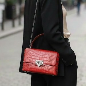 Puede incluir: Una mujer con un traje negro lleva un bolso rojo con una correa de cadena plateada. El bolso tiene una textura de efecto cocodrilo y un cierre plateado. El fondo muestra una calle de la ciudad con edificios y personas borrosas.