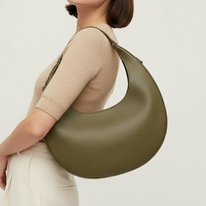 May include: Olive green crescent-shaped shoulder bag with a textured surface. The bag has a long strap and a curved design. The bag is worn over the shoulder. The bag is a neutral accessory.