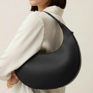 May include: Black crescent-shaped handbag with a textured surface. The bag has a single strap and a curved design. The bag is held by a person wearing a white button-down shirt.