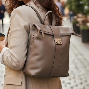 May include: A woman wearing a beige blazer and a brown leather backpack with a top handle and a flap closure. She is holding a takeaway coffee cup. The background shows a cobbled street and buildings.