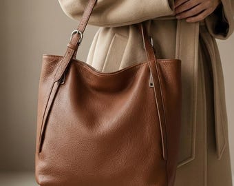 Peut inclure: Un sac fourre-tout en cuir marron avec une bandoulière. Le sac a un design simple avec une surface texturée et une boucle de couleur argentée. Le sac est tenu par une personne portant un manteau beige.