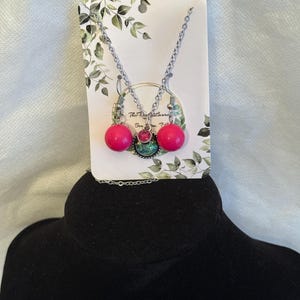 May include: A jewelry set featuring a silver-tone chain necklace and matching drop earrings on a display card. The earrings have bright pink spherical beads. The necklace pendant includes a small pink rhinestone and a larger iridescent green cabochon.