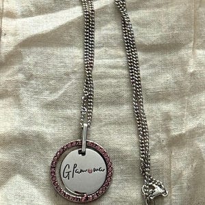 May include: Silver-toned necklace with a circular pendant. The pendant has a pink gemstone border and the handwritten text "Glam-ma" in black. The necklace chain is a curb style with a lobster clasp.