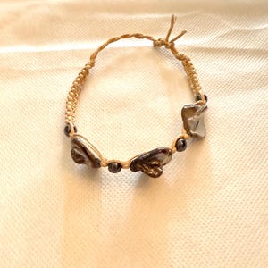 May include: A handmade bracelet featuring a natural, woven cord. The bracelet is adorned with three irregularly shaped, iridescent shell pieces and small, dark beads. The cord is tied at the ends to adjust the size.