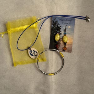 May include: A jewelry set featuring a necklace, earrings, and a bracelet. The necklace has a blue cord and a white pendant with a blue design. The earrings and necklace have yellow oval beads. The bracelet is silver with yellow beads.