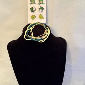 May include: A black jewelry display stand holds a beaded necklace and a set of earrings. The earrings are on a white card and feature green leprechaun hats, shamrocks, and leaves. The necklace is made of yellow, green, and gold beads.