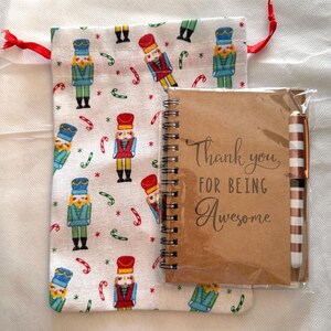 May include: A gift set featuring a small notebook, pen, and drawstring bag. The notebook has the text "Thank you, FOR BEING Awesome". The bag is white with a pattern of nutcrackers and candy canes. The pen has a striped design.