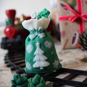 Può includere: Una borsa verde a tema natalizio con fiocchi di neve bianchi e un disegno di albero di Natale. La borsa è su un binario di treno in miniatura, con due piante grasse verdi davanti. Un trenino rosso e nero è sullo sfondo.