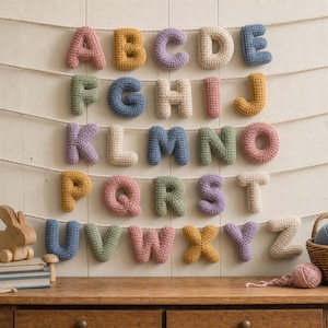 Puede incluir: Un conjunto de veintiséis letras de ganchillo de la A a la Z, cada una elaborada en varios colores de hilo pastel y apagados. Las letras están suspendidas en múltiples cuerdas blancas horizontales, creando un alfabeto colgado en la pared.