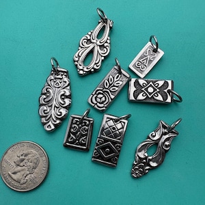 Puede incluir: Una colecci&oacute;n de ocho dijes de metal en tono plateado con varios patrones en relieve, incluyendo dise&ntilde;os florales, geom&eacute;tricos y de volutas, cada uno con una peque&ntilde;a argolla, exhibidos junto a una moneda de veinticinco centavos de EE. UU.