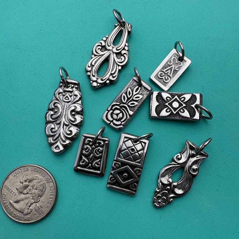 Puede incluir: Una colecci&oacute;n de ocho dijes de metal en tono plateado con varios patrones en relieve, incluyendo dise&ntilde;os florales, geom&eacute;tricos y de volutas, cada uno con una peque&ntilde;a argolla, exhibidos junto a una moneda de veinticinco centavos de EE. UU.