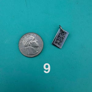 Puede incluir: Un colgante rectangular de metal plateado con un dise&ntilde;o en relieve de una letra A estilizada con volutas decorativas, colocado junto a una moneda de 25 centavos de d&oacute;lar estadounidense para comparar el tama&ntilde;o, con el n&uacute;mero 9 impreso debajo.