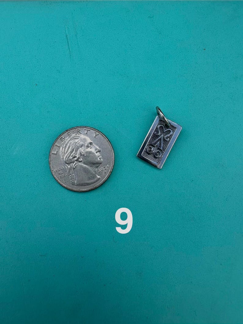 Puede incluir: Un colgante rectangular de metal plateado con un dise&ntilde;o en relieve de una letra A estilizada con volutas decorativas, colocado junto a una moneda de 25 centavos de d&oacute;lar estadounidense para comparar el tama&ntilde;o, con el n&uacute;mero 9 impreso debajo.