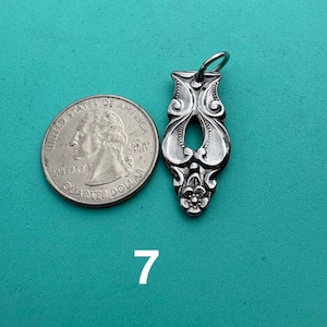 Puede incluir: Un colgante de metal plateado con un patr&oacute;n floral ornamentado y alargado, una abertura central y una peque&ntilde;a anilla en la parte superior, colocado junto a una moneda de 25 centavos de EE. UU. para escala, con el n&uacute;mero 7 impreso debajo.