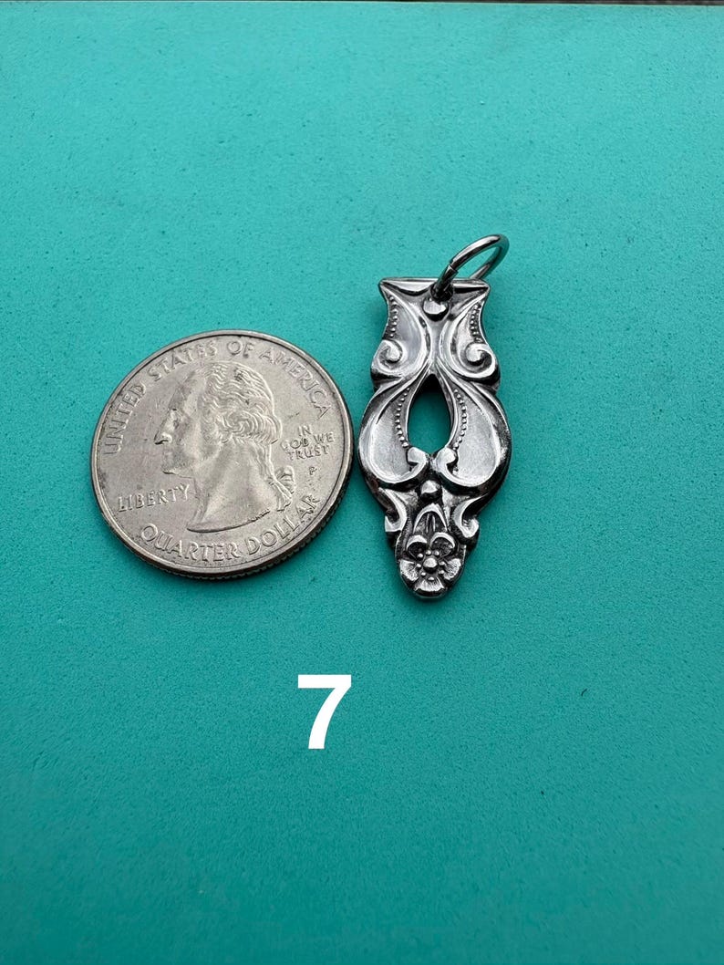 Puede incluir: Un colgante de metal plateado con un patr&oacute;n floral ornamentado y alargado, una abertura central y una peque&ntilde;a anilla en la parte superior, colocado junto a una moneda de 25 centavos de EE. UU. para escala, con el n&uacute;mero 7 impreso debajo.