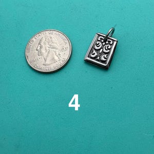 Puede incluir: Un colgante rectangular de plata con un patr&oacute;n de volutas en relieve y una peque&ntilde;a argolla en la parte superior, colocado junto a una moneda de 25 centavos de EE. UU. para escala. El n&uacute;mero 4 est&aacute; impreso en blanco debajo del colgante.