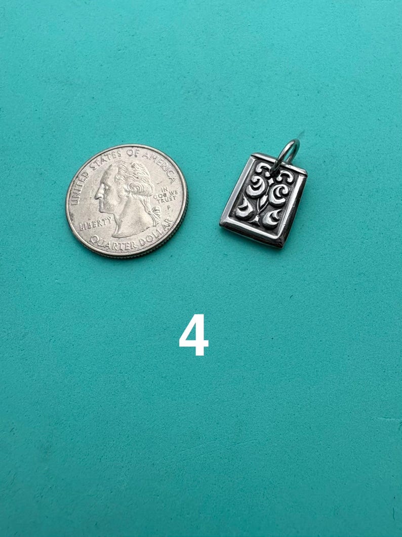 Puede incluir: Un colgante rectangular de plata con un patr&oacute;n de volutas en relieve y una peque&ntilde;a argolla en la parte superior, colocado junto a una moneda de 25 centavos de EE. UU. para escala. El n&uacute;mero 4 est&aacute; impreso en blanco debajo del colgante.