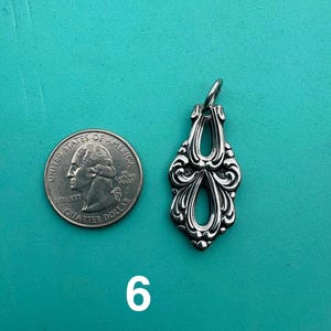 Puede incluir: Un colgante de metal plateado con un dise&ntilde;o ornamental sim&eacute;trico y dos aberturas. El colgante est&aacute; colocado junto a una moneda de veinticinco centavos de d&oacute;lar de los Estados Unidos para mostrar la escala, y el n&uacute;mero 6 aparece debajo.