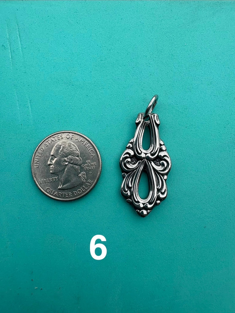Puede incluir: Un colgante de metal plateado con un dise&ntilde;o ornamental sim&eacute;trico y dos aberturas. El colgante est&aacute; colocado junto a una moneda de veinticinco centavos de d&oacute;lar de los Estados Unidos para mostrar la escala, y el n&uacute;mero 6 aparece debajo.