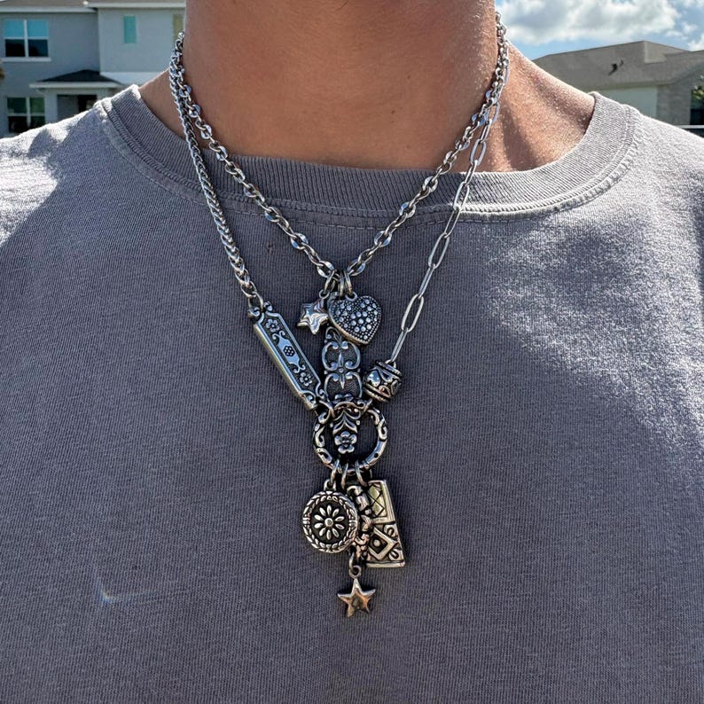 Puede incluir: Un collar de capas en tono plateado con dos cadenas distintas. La cadena superior es de estilo cl&aacute;sico, mientras que la cadena inferior presenta un dise&ntilde;o de eslabones mixtos adornado con m&uacute;ltiples dijes, incluyendo un coraz&oacute;n, una estrella y una flor.