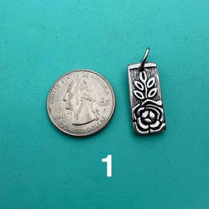 Puede incluir: Un colgante rectangular de metal plateado con un dise&ntilde;o en relieve de una rosa y hojas, colocado junto a una moneda de 25 centavos de EE. UU. para comparar el tama&ntilde;o. El colgante tiene una peque&ntilde;a anilla superior y una superficie texturizada.