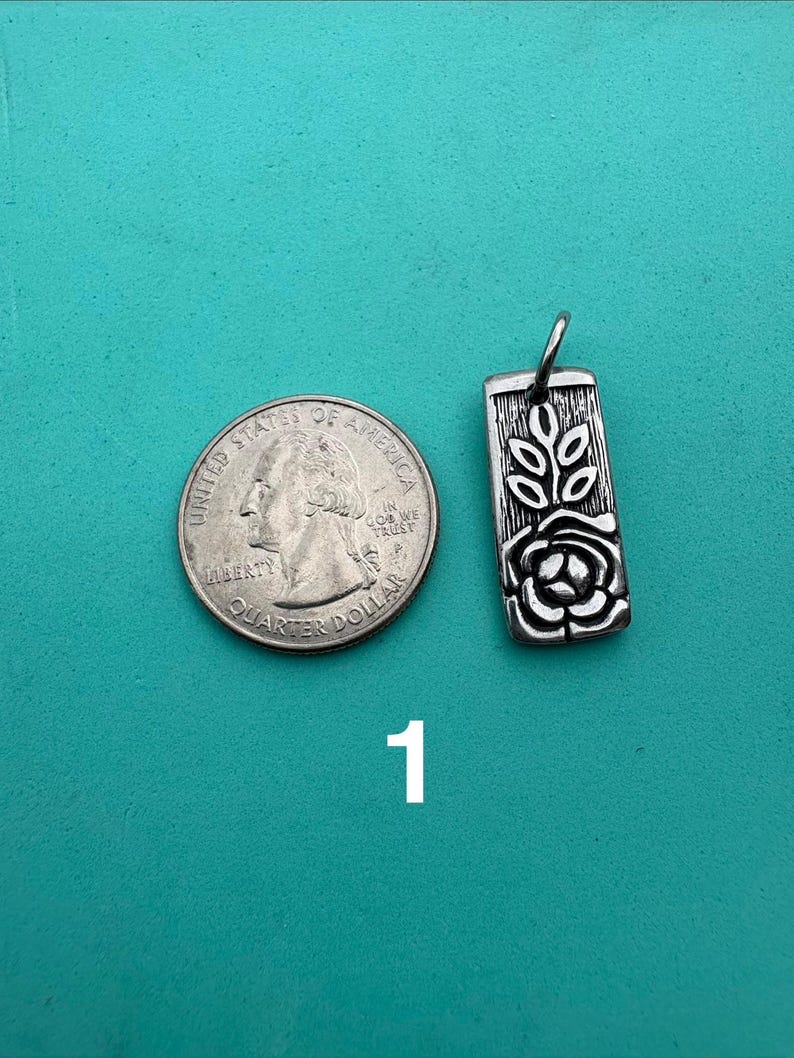 Puede incluir: Un colgante rectangular de metal plateado con un dise&ntilde;o en relieve de una rosa y hojas, colocado junto a una moneda de 25 centavos de EE. UU. para comparar el tama&ntilde;o. El colgante tiene una peque&ntilde;a anilla superior y una superficie texturizada.