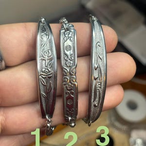 May include: Three silver-toned pendants with intricate designs. The first features a rose, the second has floral patterns, and the third has a curved shape. Each pendant is connected to a delicate chain. Numbers 1, 2, and 3 are printed below the pendants.