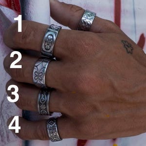 May include: Four silver-toned metal rings worn on fingers, labeled 1 through 4. Ring 1 features a central oval with a floral motif. Ring 2 displays a repeating four-petal pattern. Ring 3 shows a swirl and star design. Ring 4 has a geometric grid pattern.