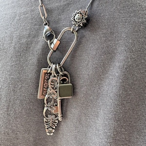 Puede incluir: Un collar de cadena plateada con un gran cierre de mosquet&oacute;n ovalado. Del cierre cuelgan varios dijes, incluyendo un peque&ntilde;o candado, una llave antigua ornamentada con detalles florales, un dije rectangular con motivos de flores y un dije de sol.