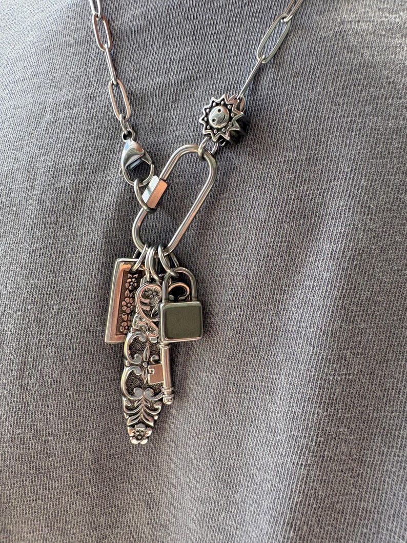 Puede incluir: Un collar de cadena plateada con un gran cierre de mosquet&oacute;n ovalado. Del cierre cuelgan varios dijes, incluyendo un peque&ntilde;o candado, una llave antigua ornamentada con detalles florales, un dije rectangular con motivos de flores y un dije de sol.
