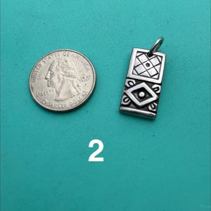 Puede incluir: Un colgante rectangular de plata con un patr&oacute;n geom&eacute;trico grabado que presenta una cuadr&iacute;cula cuadrada en la parte superior y una forma de diamante en la parte inferior, junto a una moneda de cuarto de d&oacute;lar estadounidense. El n&uacute;mero 2 aparece debajo.