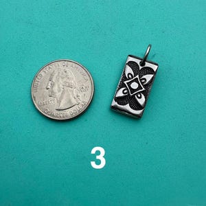 Puede incluir: Un colgante rectangular de metal plateado con un dise&ntilde;o floral sim&eacute;trico de cuatro p&eacute;talos en relieve con un punto central, colocado junto a una moneda de cuarto de d&oacute;lar estadounidense para comparar el tama&ntilde;o. El colgante incluye una peque&ntilde;a argolla.