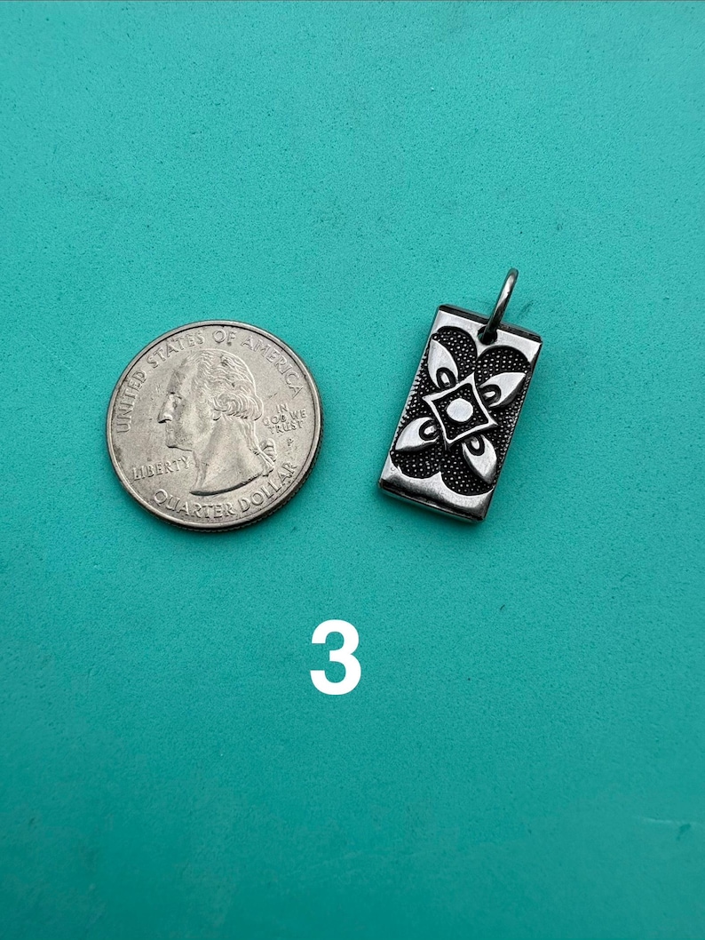 Puede incluir: Un colgante rectangular de metal plateado con un dise&ntilde;o floral sim&eacute;trico de cuatro p&eacute;talos en relieve con un punto central, colocado junto a una moneda de cuarto de d&oacute;lar estadounidense para comparar el tama&ntilde;o. El colgante incluye una peque&ntilde;a argolla.