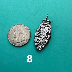 Puede incluir: Un colgante de metal plateado con forma alargada similar a una hoja, con intrincados grabados en relieve y un peque&ntilde;o lazo superior, colocado junto a una moneda de cuarto de d&oacute;lar estadounidense para escala, con el n&uacute;mero 8 impreso debajo.