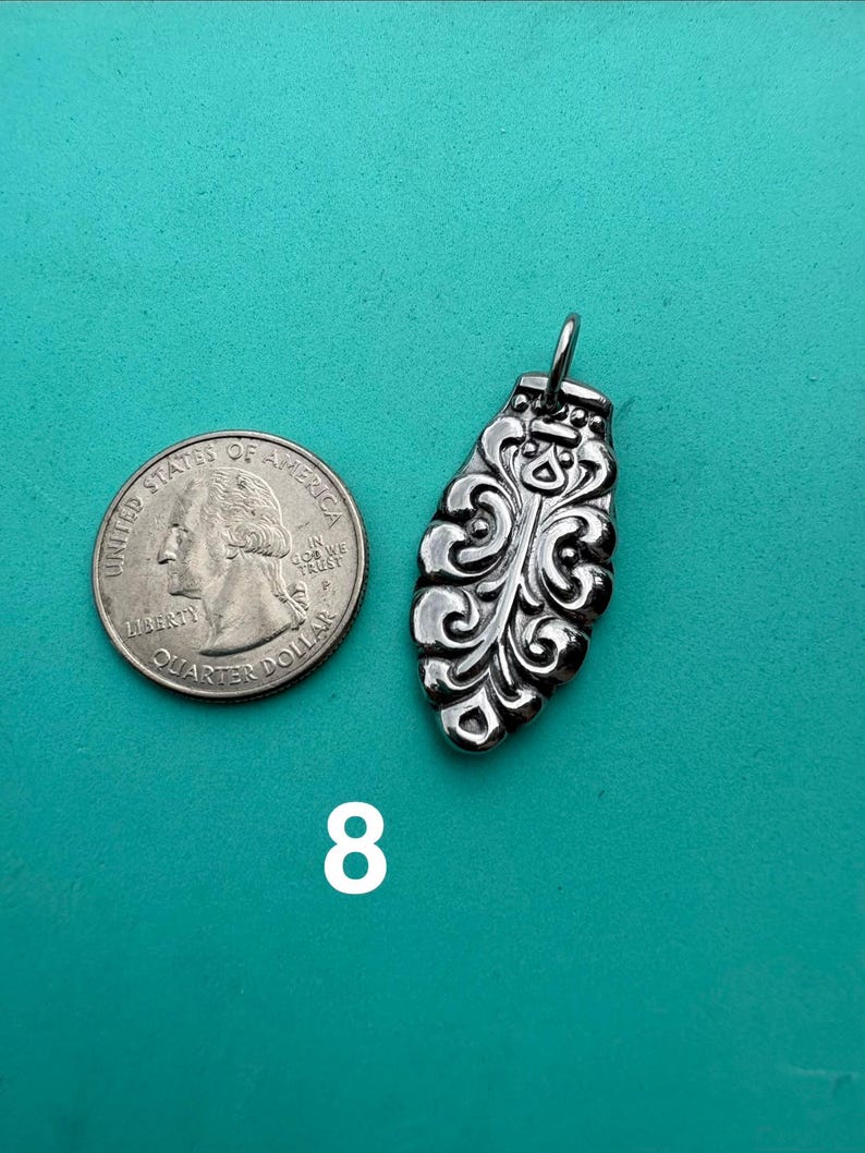 Puede incluir: Un colgante de metal plateado con forma alargada similar a una hoja, con intrincados grabados en relieve y un peque&ntilde;o lazo superior, colocado junto a una moneda de cuarto de d&oacute;lar estadounidense para escala, con el n&uacute;mero 8 impreso debajo.