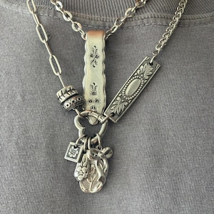 Puede incluir: Un collar plateado de varias capas con una mezcla de estilos de cadena, incluyendo eslabones ovalados largos y una cadena de bordillo. El conjunto incluye dos placas rectangulares de metal con grabados florales, una cuenta texturizada y tres dijes.