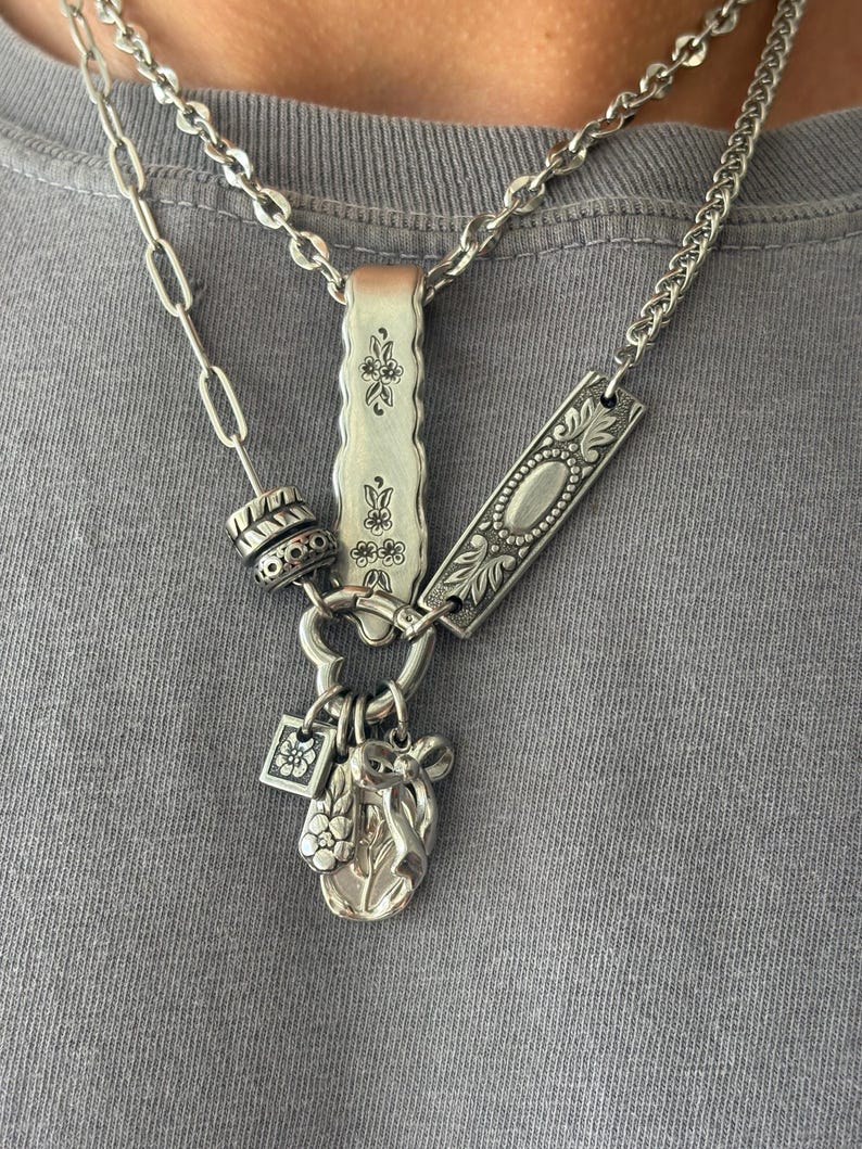 Puede incluir: Un collar plateado de varias capas con una mezcla de estilos de cadena, incluyendo eslabones ovalados largos y una cadena de bordillo. El conjunto incluye dos placas rectangulares de metal con grabados florales, una cuenta texturizada y tres dijes.