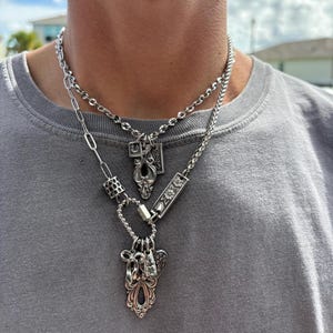 Puede incluir: Un collar de capas en tono plateado con dos cadenas distintas. La cadena superior tiene un peque&ntilde;o colgante rectangular, mientras que la inferior incorpora una mezcla de eslabones ovalados, una cuenta texturizada y un cierre de mosquet&oacute;n.