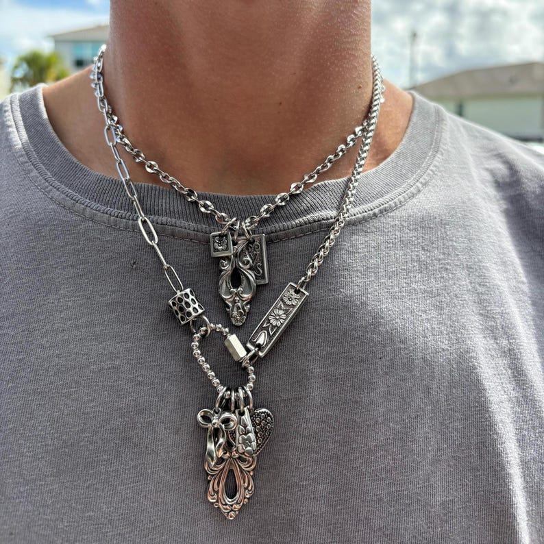 Puede incluir: Un collar de capas en tono plateado con dos cadenas distintas. La cadena superior tiene un peque&ntilde;o colgante rectangular, mientras que la inferior incorpora una mezcla de eslabones ovalados, una cuenta texturizada y un cierre de mosquet&oacute;n.