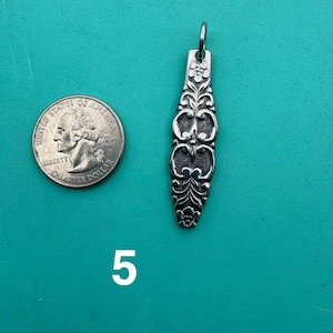 Puede incluir: Un colgante de metal plateado con un patr&oacute;n floral alargado y ornamentado, con una peque&ntilde;a anilla en la parte superior. El colgante est&aacute; colocado junto a una moneda de cuarto de d&oacute;lar de EE. UU. para comparar el tama&ntilde;o, y el n&uacute;mero 5 aparece impreso debajo.