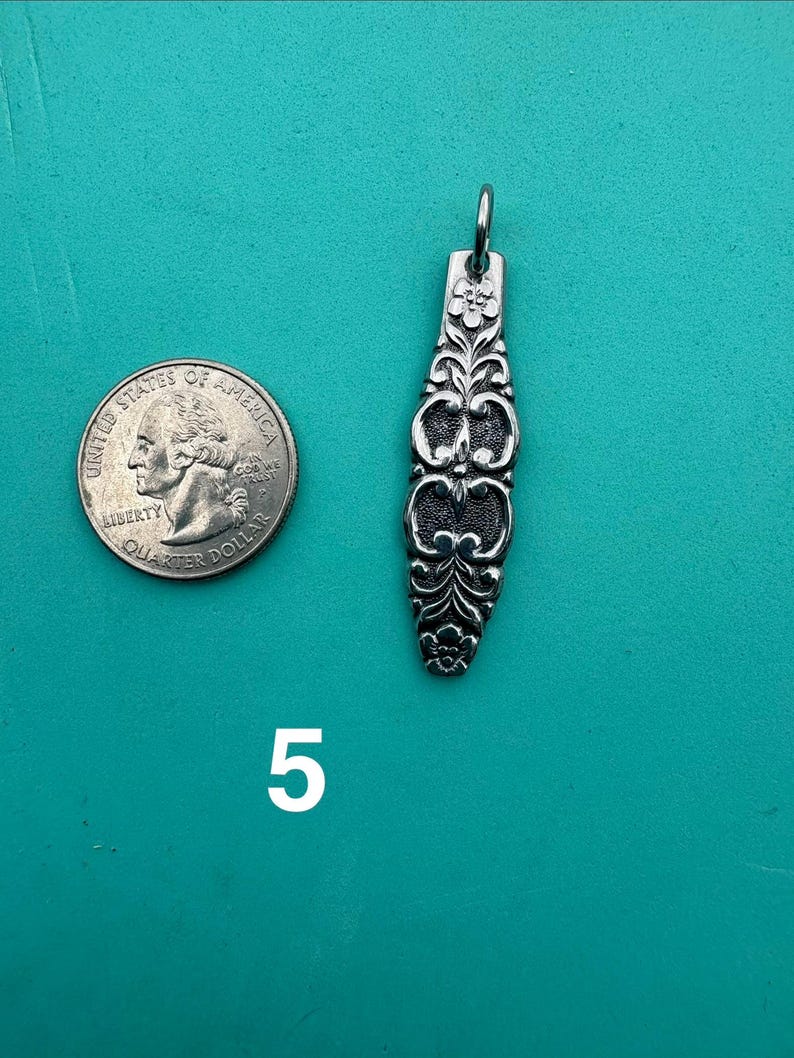 Puede incluir: Un colgante de metal plateado con un patr&oacute;n floral alargado y ornamentado, con una peque&ntilde;a anilla en la parte superior. El colgante est&aacute; colocado junto a una moneda de cuarto de d&oacute;lar de EE. UU. para comparar el tama&ntilde;o, y el n&uacute;mero 5 aparece impreso debajo.