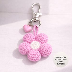 May include: A pink crochet flower keyring with a silver clasp and a small pink heart charm. The flower has a white centre and six pink petals. The keyring is a handmade craft item.