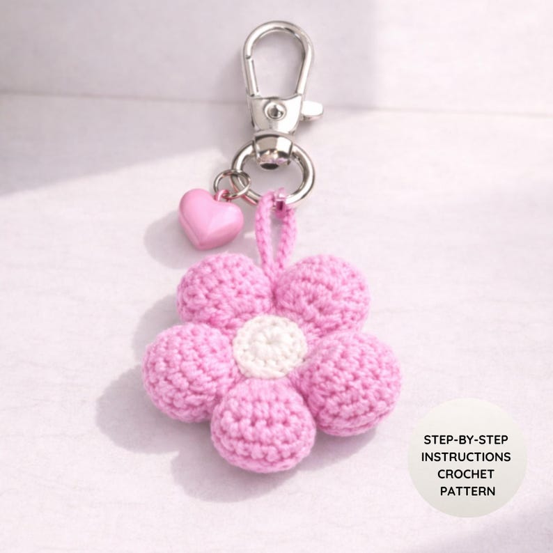 May include: A pink crochet flower keyring with a silver clasp and a small pink heart charm. The flower has a white centre and six pink petals. The keyring is a handmade craft item.