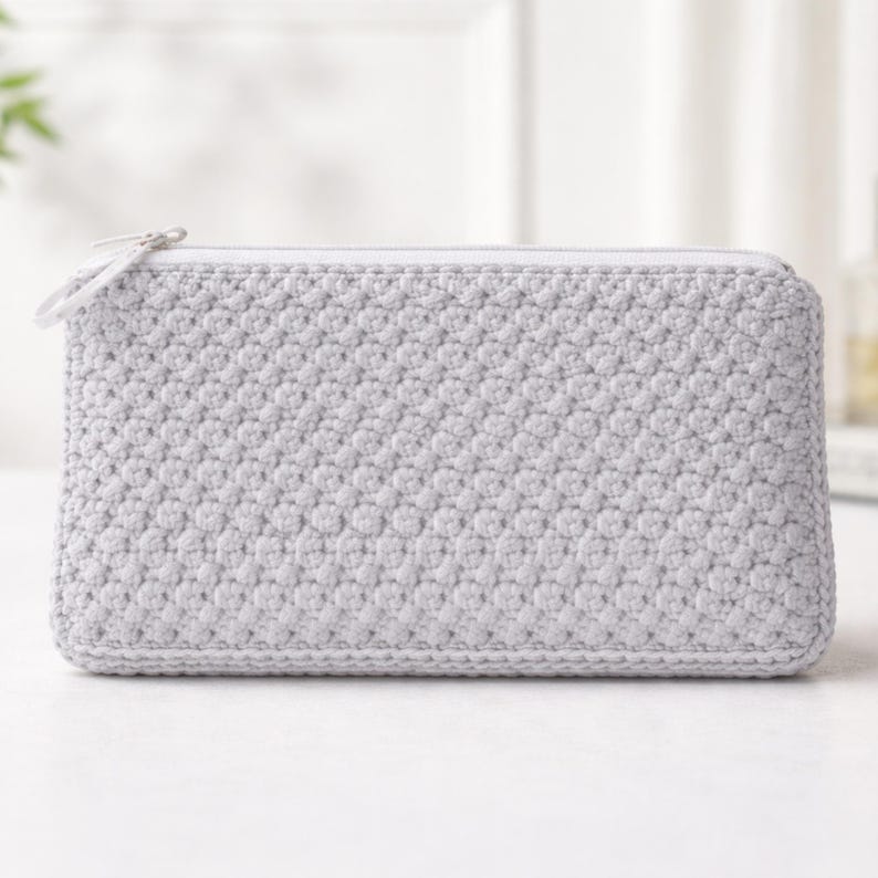 K&ouml;nnte beinhalten: Eine hellgraue, rechteckige H&auml;keltasche mit Rei&szlig;verschluss. Die Tasche hat ein strukturiertes, kn&ouml;tchenartiges Muster und einen wei&szlig;en Rei&szlig;verschlussanh&auml;nger. Das Design ist schlicht und minimalistisch, geeignet f&uuml;r kleine Gegenst&auml;nde.