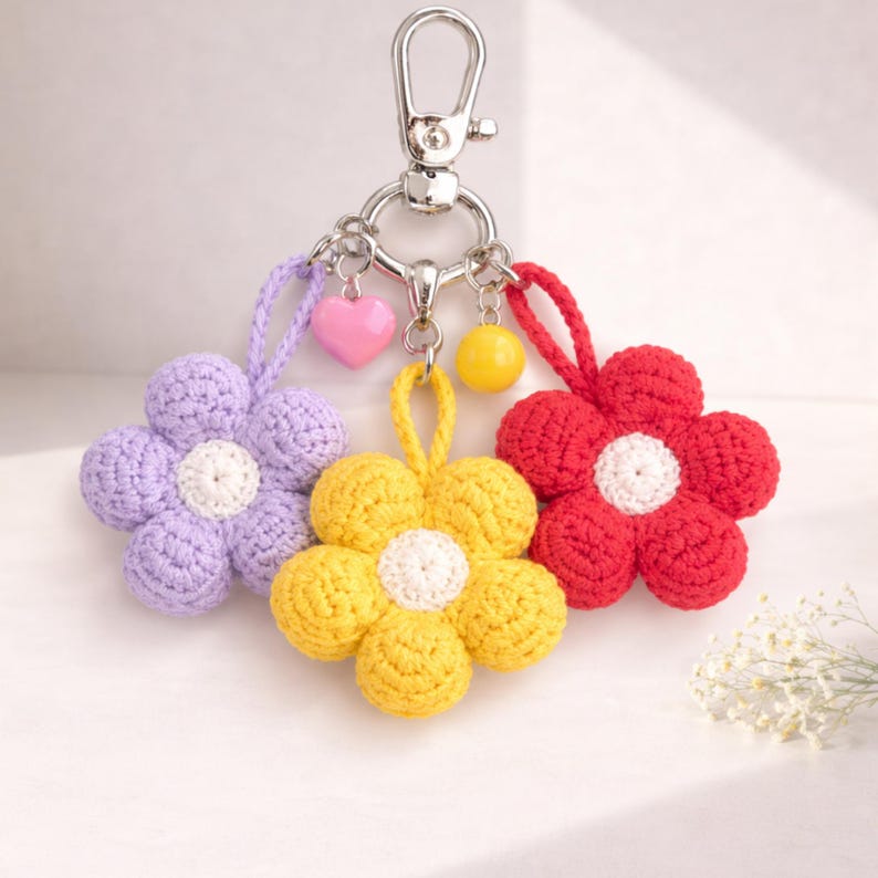 May include: A keychain featuring three crochet flower charms in purple, yellow, and red. Each flower has a white centre and is attached to a silver metal ring with a lobster clasp. Small charms include a pink heart and a yellow ball.