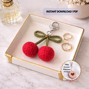 Puede incluir: Un llavero de cerezas rojas de ganchillo hecho a mano, con tallo y lazo verdes, en una bandeja blanca con borde dorado. La imagen incluye el texto "INSTANT DOWNLOAD | PDF" y "FREE Cookie Keychain Pattern".