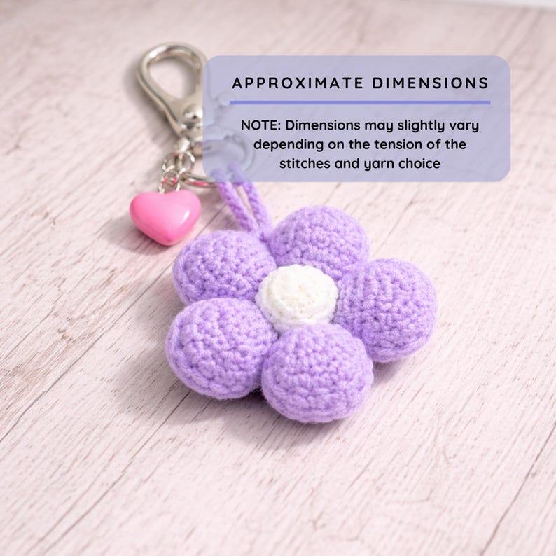 May include: A lilac and white crochet flower keyring. The flower has six petals and a white centre, attached to a silver-tone clasp with a pink heart charm. The text "APPROXIMATE DIMENSIONS" is visible.