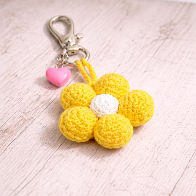 May include: A yellow crochet flower keyring with a white centre and a pink heart charm. The keyring has a silver clasp for attaching to keys or bags. The flower is made of soft yarn.