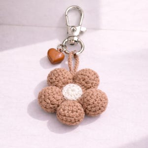 May include: A handmade keychain with a crocheted flower design. The flower has five brown petals and a white centre. A small brown heart charm hangs from the silver clasp. Ideal for keys or bags.